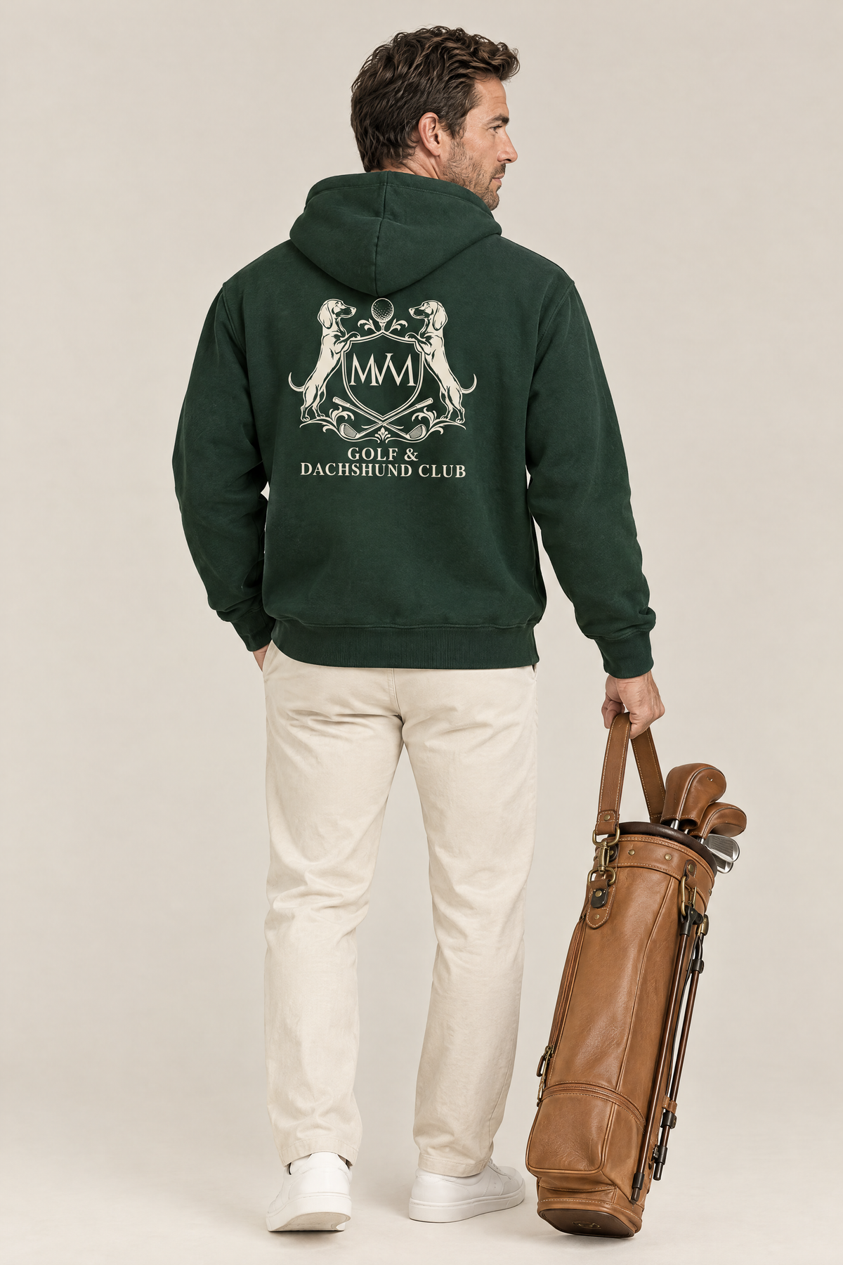 Man carrying a tan leather golf bag, back view of the blackish green Club Crest hoodie with the large cream crest. Color: Blackish Green