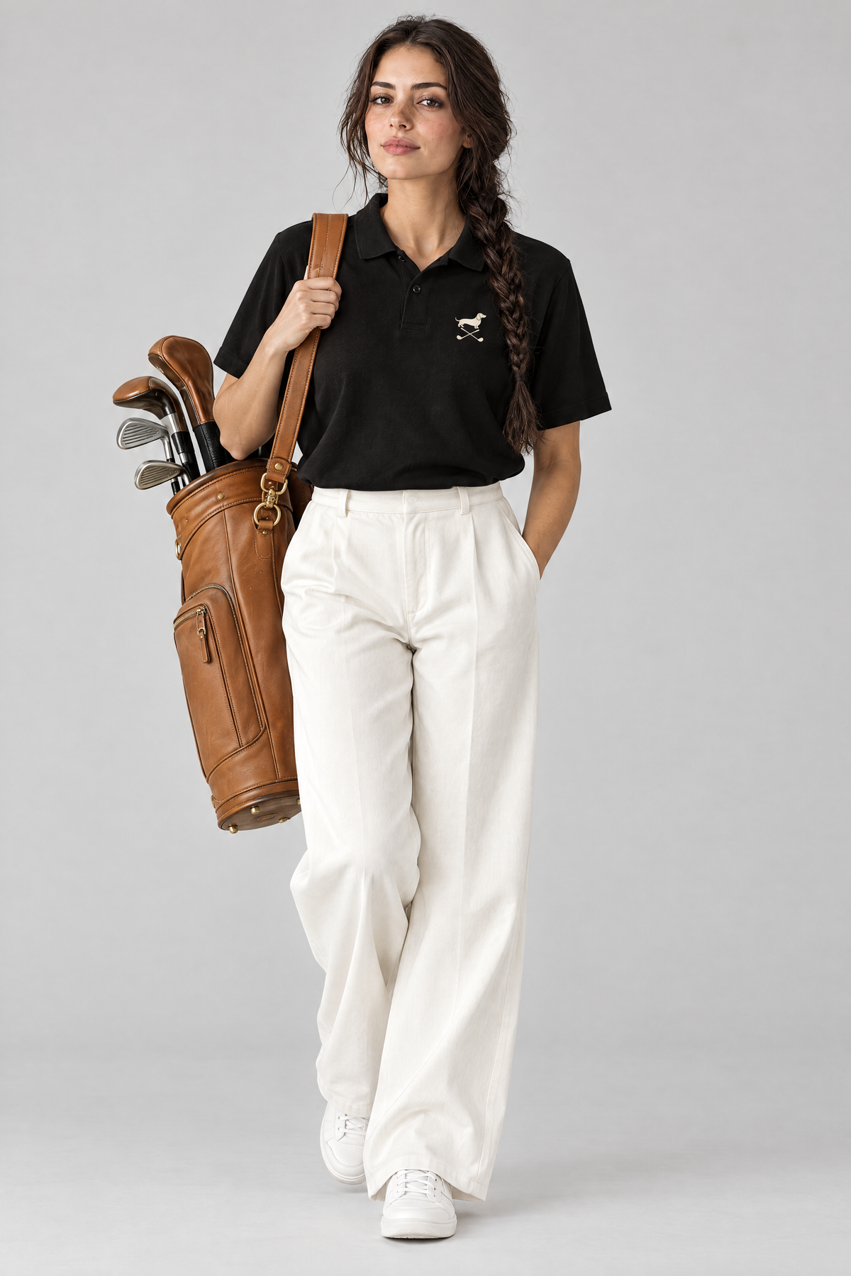 Woman carrying a tan leather golf bag in the black Club Mark dachshund golf polo and white wide-leg trousers. Color: Black
