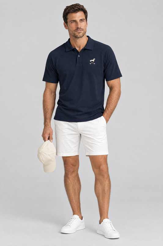 Man holding a cream golf cap in the navy blue Club Mark dachshund golf polo and white golf shorts. Color: Navy Blue