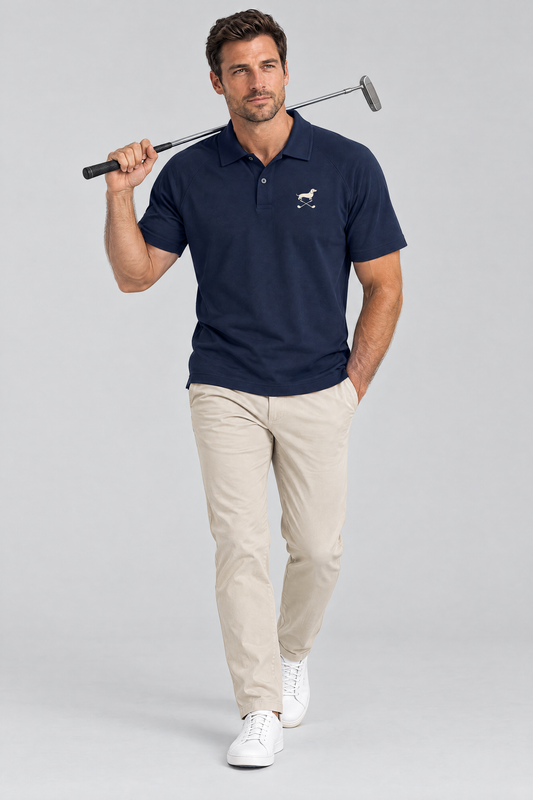 Man walking with a putter on his shoulder in the navy blue Club Mark dachshund golf polo and stone chinos. Color: Navy Blue