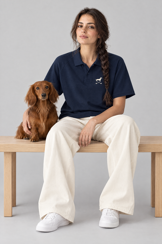 Woman sitting on a wooden bench with a long-haired dachshund in the navy blue Club Mark dachshund golf polo and cream wide-leg trousers. Color: Navy Blue
