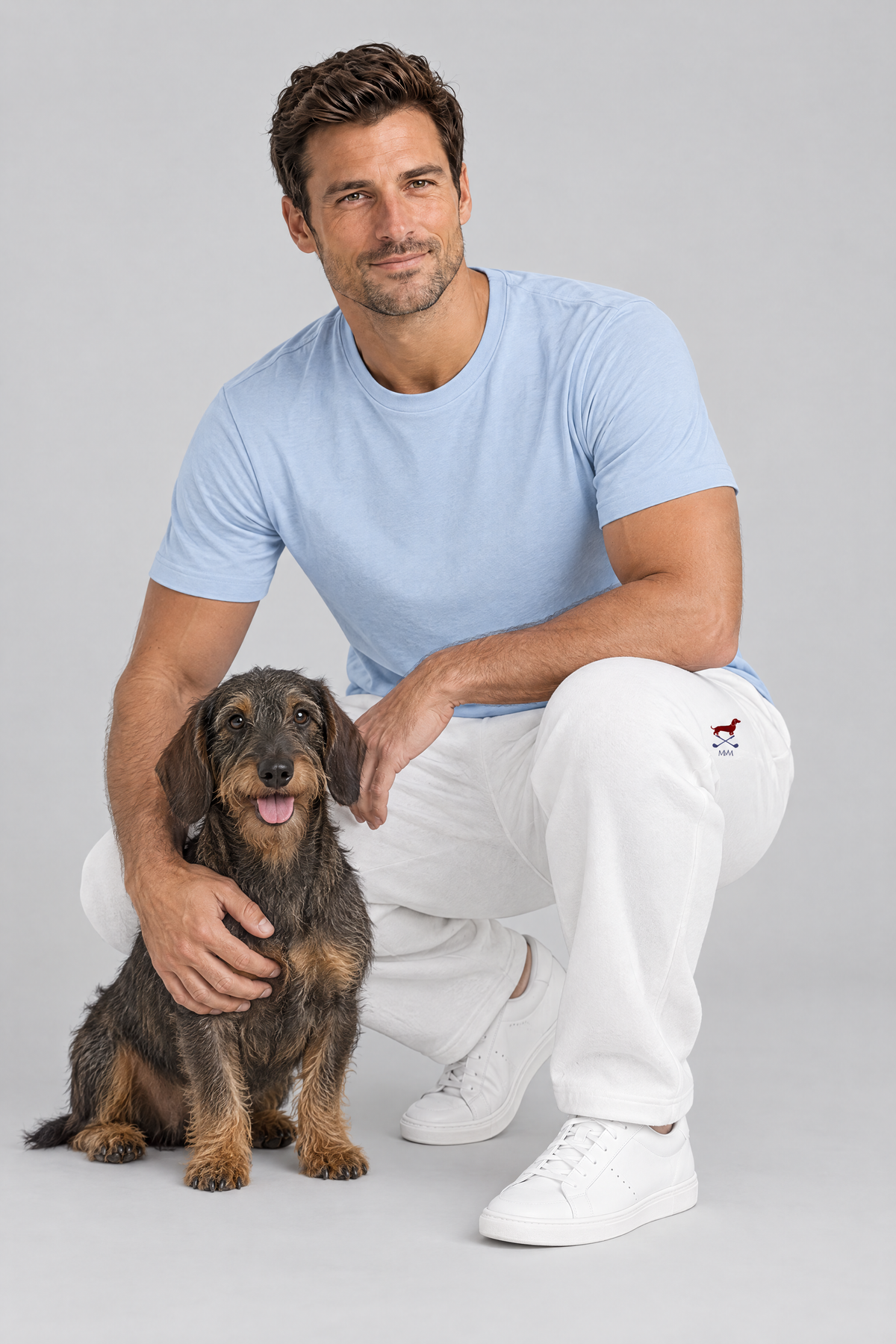 Man crouching next to a wire-haired dachshund in white Club Mark dachshund golf sweatpants and a light blue tee. Color: White