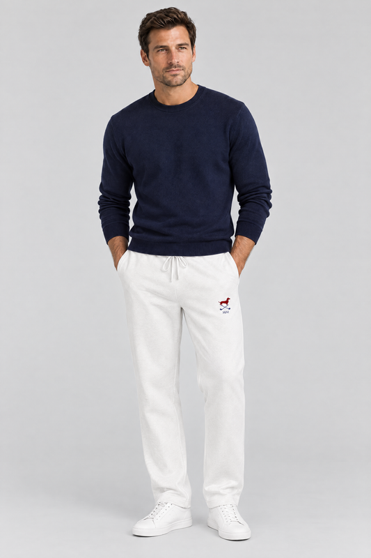 Man standing with his hands in his pockets in white Club Mark dachshund golf sweatpants with the small red dachshund logo and a navy sweater. Color: White