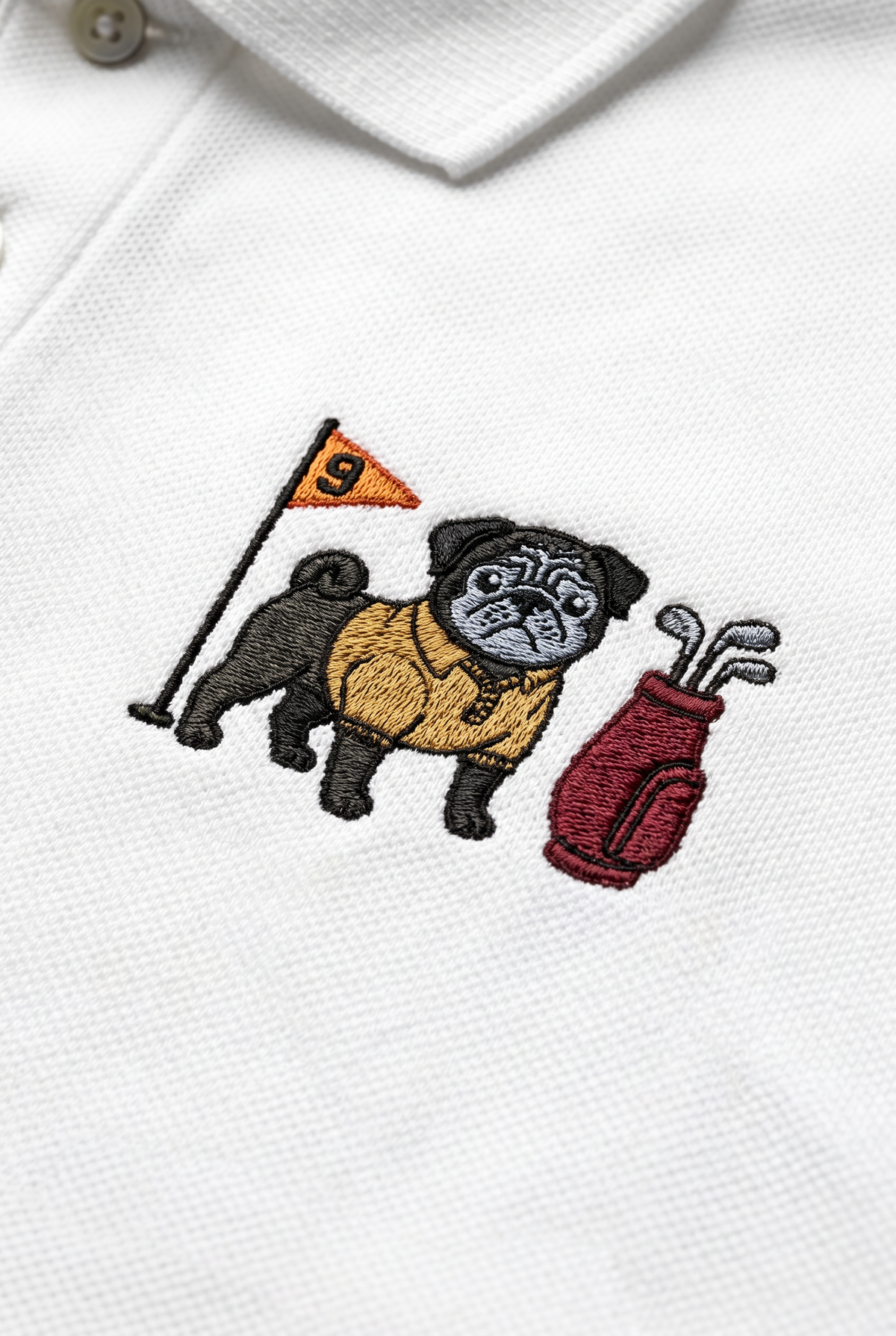 Close-up of a white polo shirt featuring an embroidered design of a pug wearing a yellow sweater, holding a golf flag next to a red golf bag.