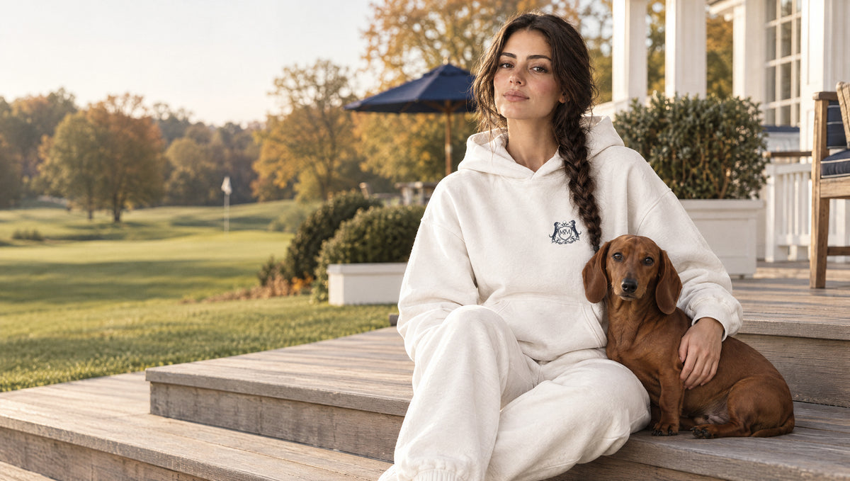 Woman in the white MVM Club Crest hoodie and sweatpants sitting with her dachshund on a golf clubhouse terrace