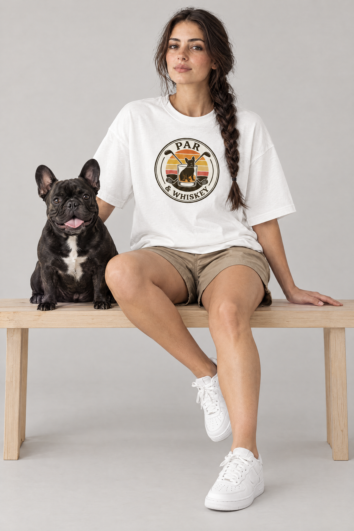 Woman sitting on a bench with a black French bulldog in the white Par & Whiskey French Bulldog Badge Golf Tee. Color: White