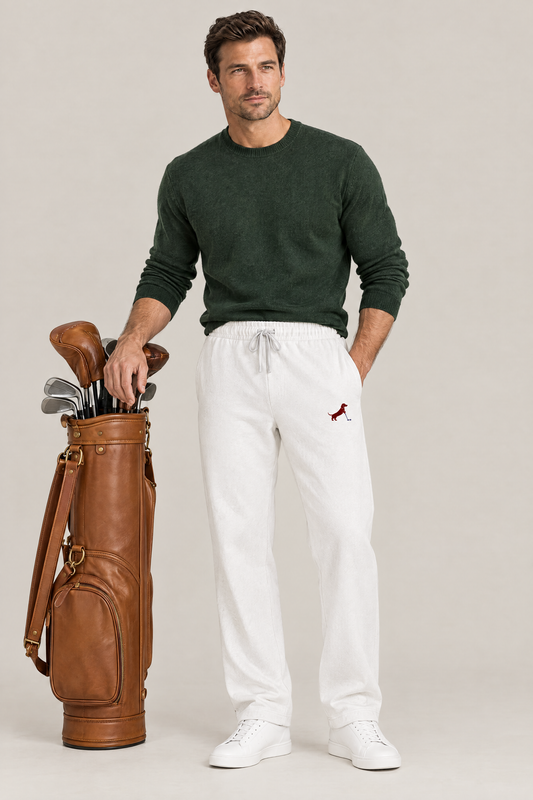Man standing next to a tan leather golf bag in white Putting dachshund golf sweatpants with the small burgundy dachshund print on the thigh and a dark green knit sweater. Color: White