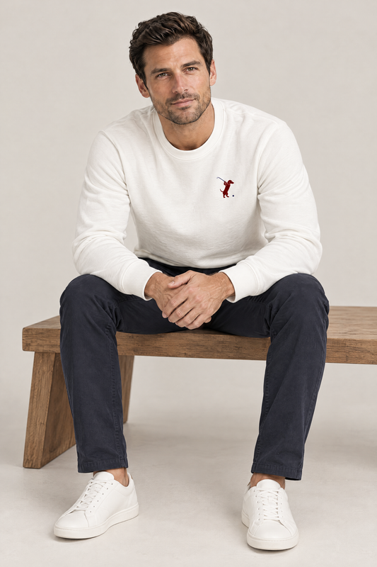 Man sitting on a low wooden bench leaning forward in white Swing dachshund golf sweatshirt and navy chinos. Color: White