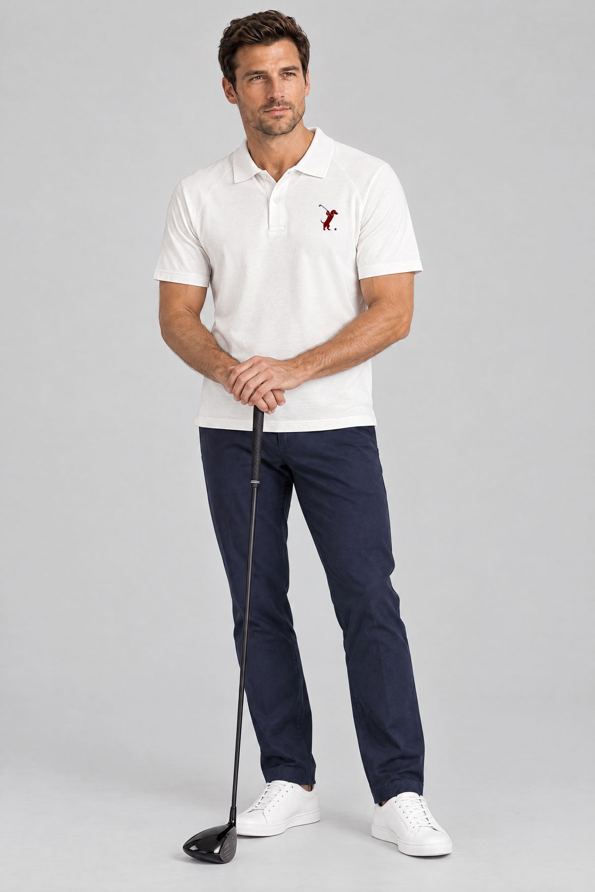 Man resting both hands on a driver in the white Swing dachshund golf polo and navy trousers. Color: White