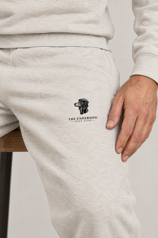 Man wearing the sweatpants, close-up of The Underdog Golf Club dachshund crest on the left thigh of the Oat Gray sweatpants. Color: Oat Gray