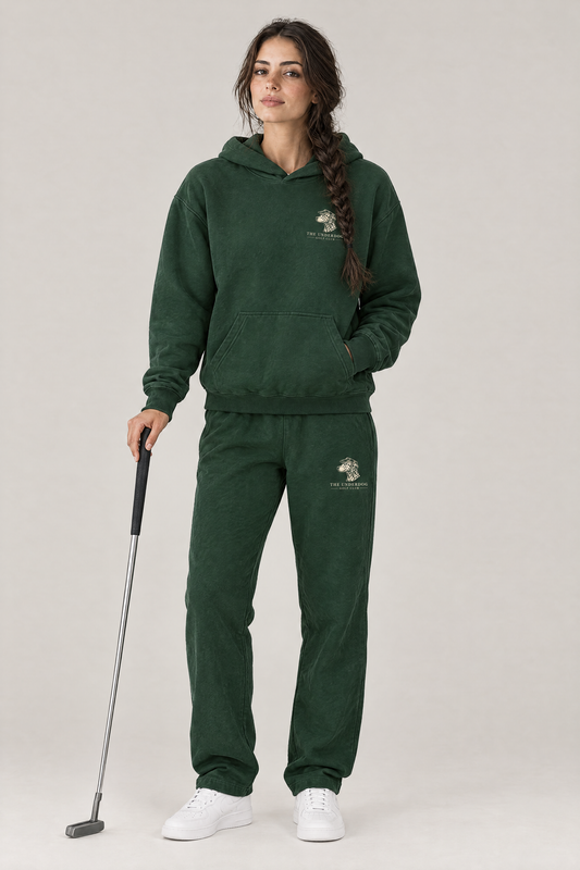 Woman holding a putter in The Sweet Spot Dalmatian golf hoodie and matching Underdog Golf Club sweatpants in Blackish Green, front view. Color: Blackish Green