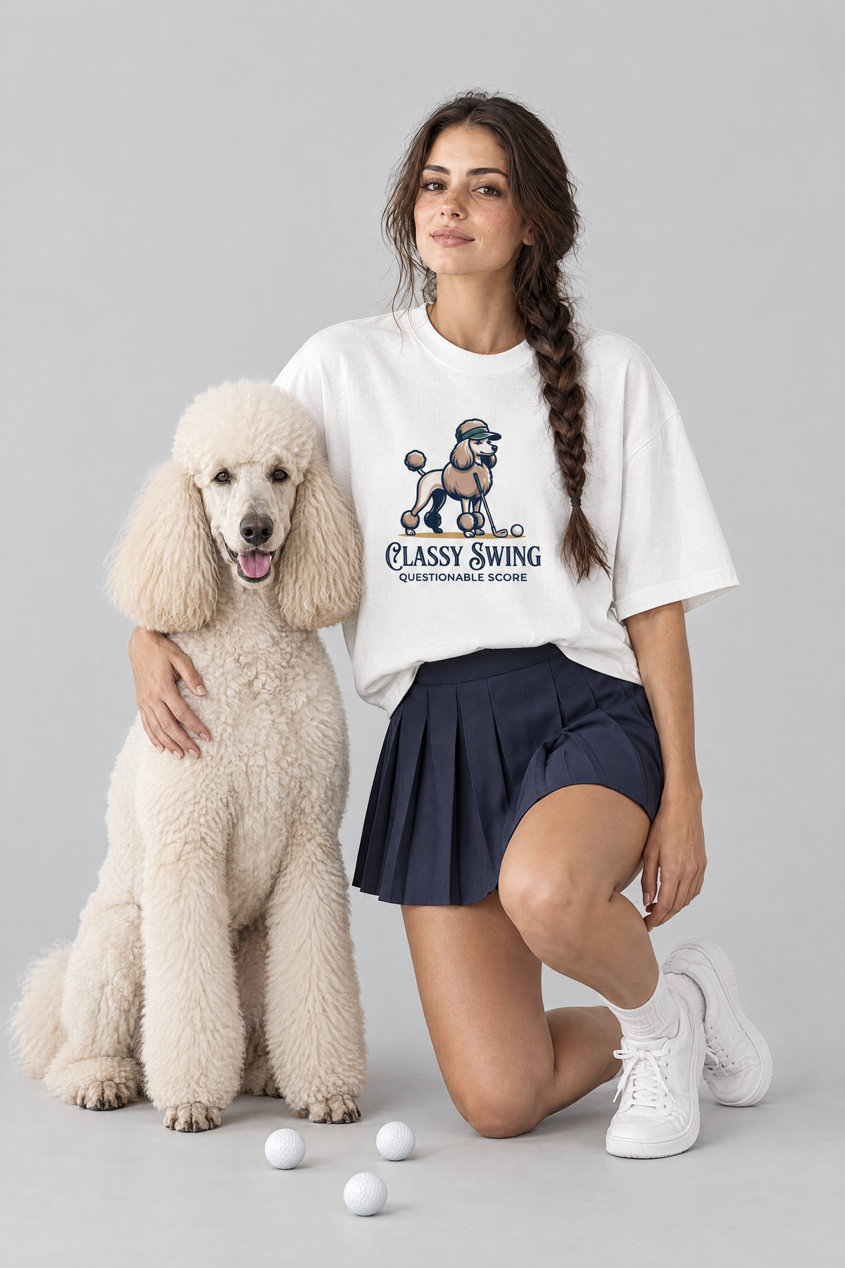 Woman kneeling beside a cream standard poodle, wearing the Classy Swing Poodle Golf Tee. Color: White