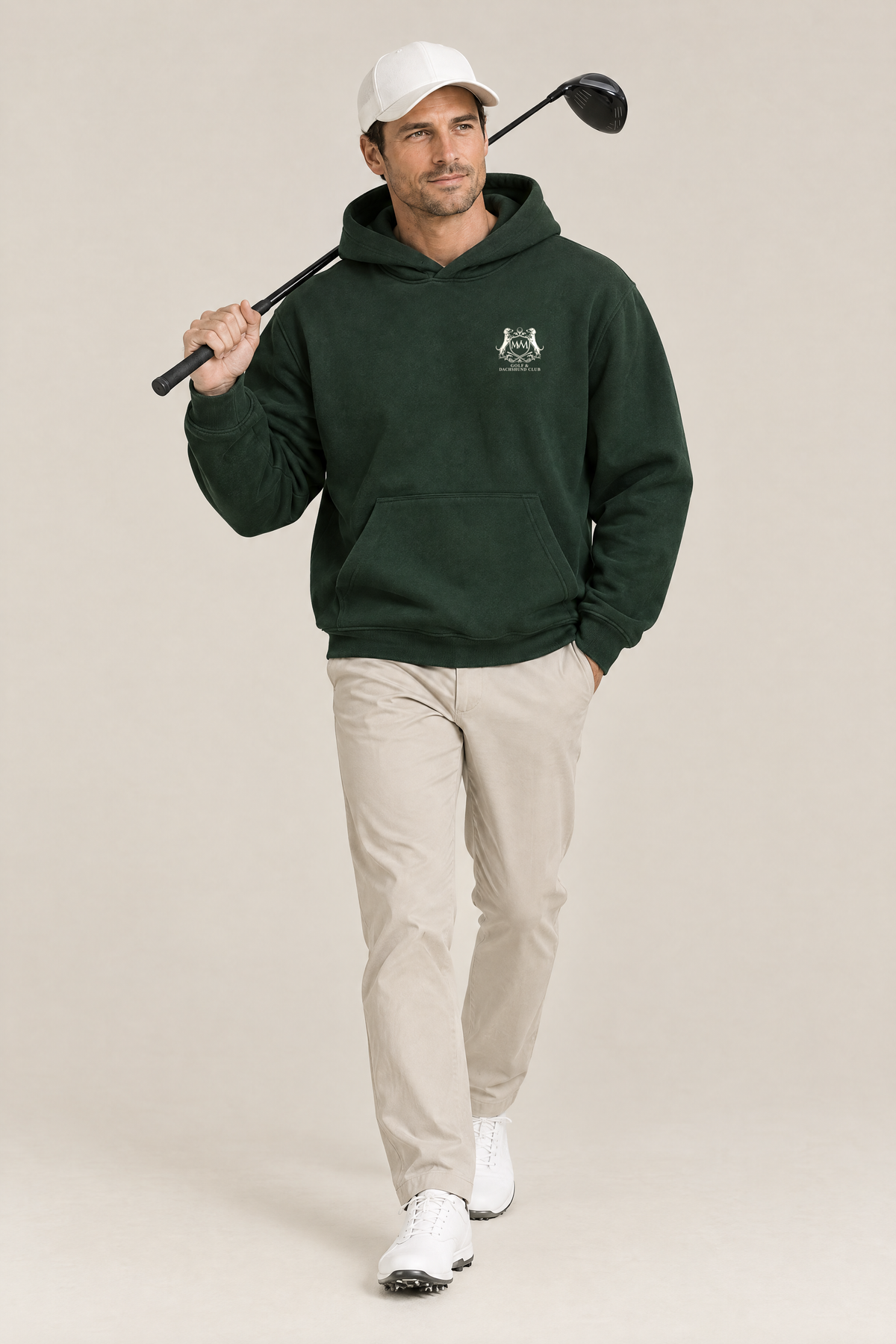 Man walking with a driver over his shoulder, wearing the blackish green Club Crest hoodie and a white cap. Color: Blackish Green