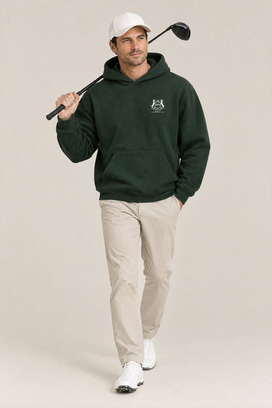 Man walking with a driver over his shoulder, wearing the blackish green Club Crest hoodie and a white cap. Color: Blackish Green