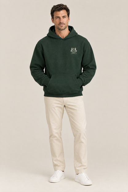 Man wearing the Club Crest hoodie in blackish green with cream chinos, hands in the front pocket. Color: Blackish Green