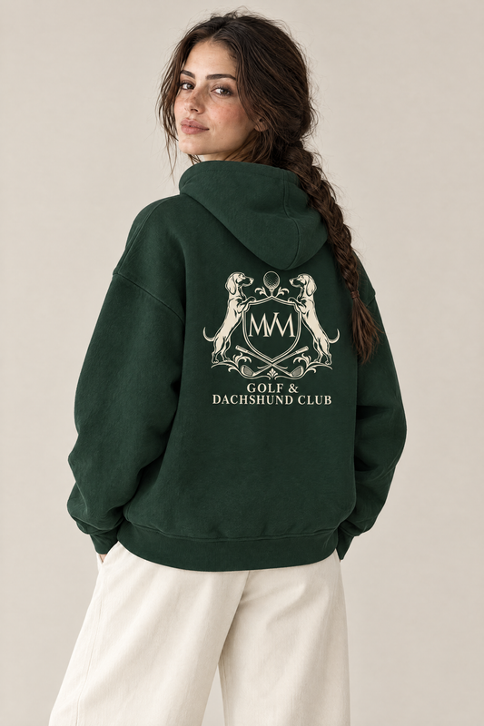 Woman looking over her shoulder, showing the large cream Golf & Dachshund Club crest on the back of the blackish green Club Crest hoodie. Color: Blackish Green