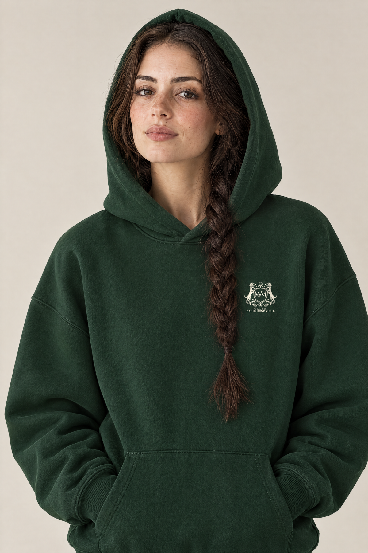 Woman wearing the Club Crest hoodie in blackish green with the hood up and the small cream Golf & Dachshund Club crest on the left chest. Color: Blackish Green