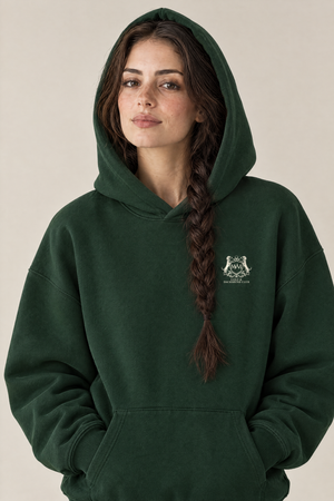 Woman wearing the Club Crest hoodie in blackish green with the hood up and the small cream Golf & Dachshund Club crest on the left chest. Color: Blackish Green