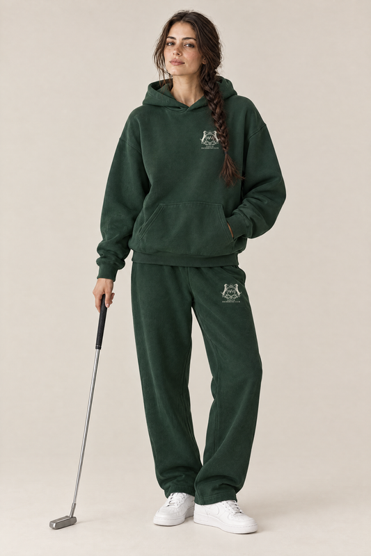 Woman in a matching blackish green set of Club Crest hoodie and sweatpants with the small cream crest, holding a putter. Color: Blackish Green