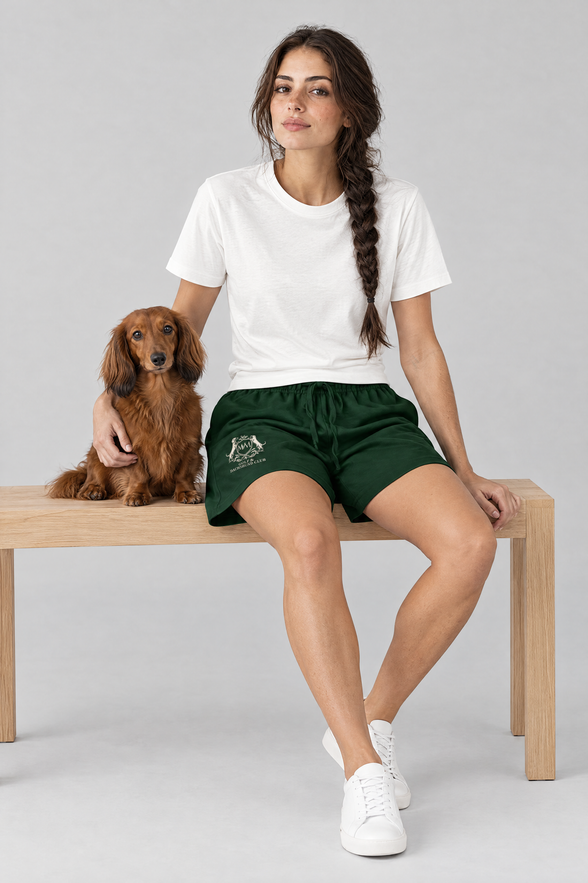 Woman sitting on a wooden bench with a long-haired dachshund, wearing blackish green Club Crest shorts and a white tee. Color: Blackish Green