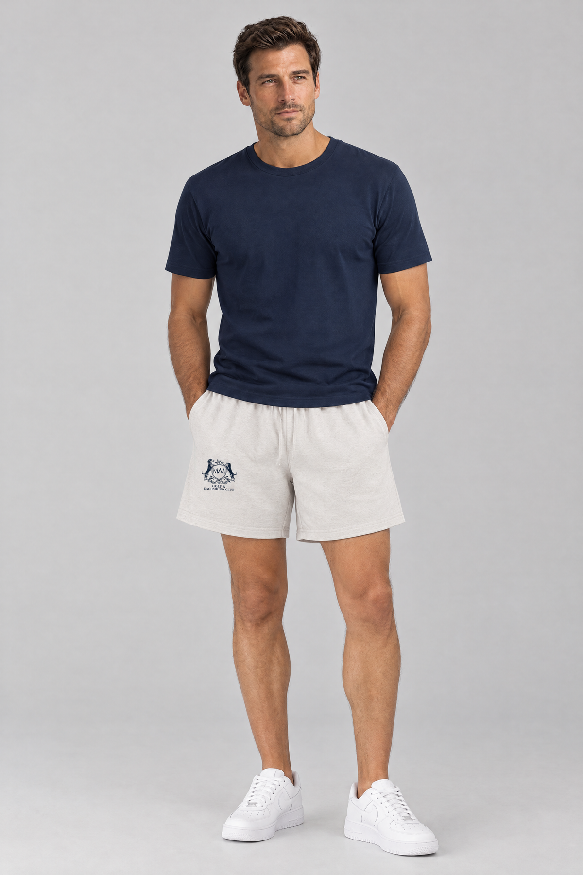 Man standing with his hands in his pockets in oat gray Club Crest shorts with the small navy crest and a navy tee. Color: Oat Gray