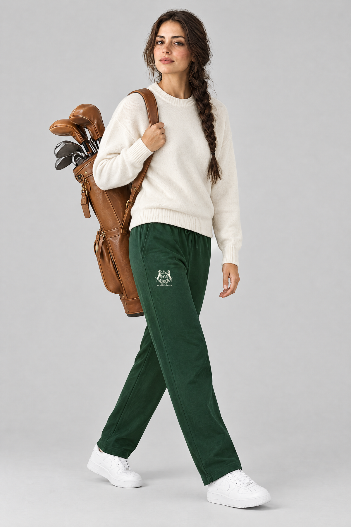 Woman walking with a tan leather golf bag in blackish green Club Crest sweatpants and a cream knit sweater. Color: Blackish Green