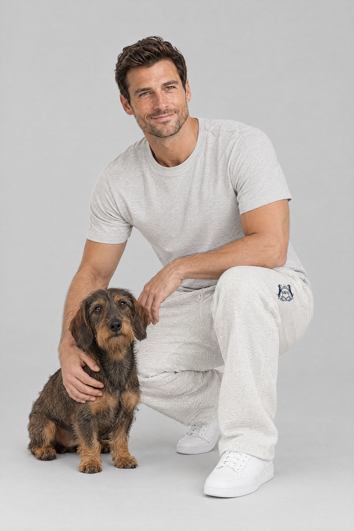 Man crouching next to a wire-haired dachshund in oat gray Club Crest sweatpants and a light grey tee. Color: Oat Gray