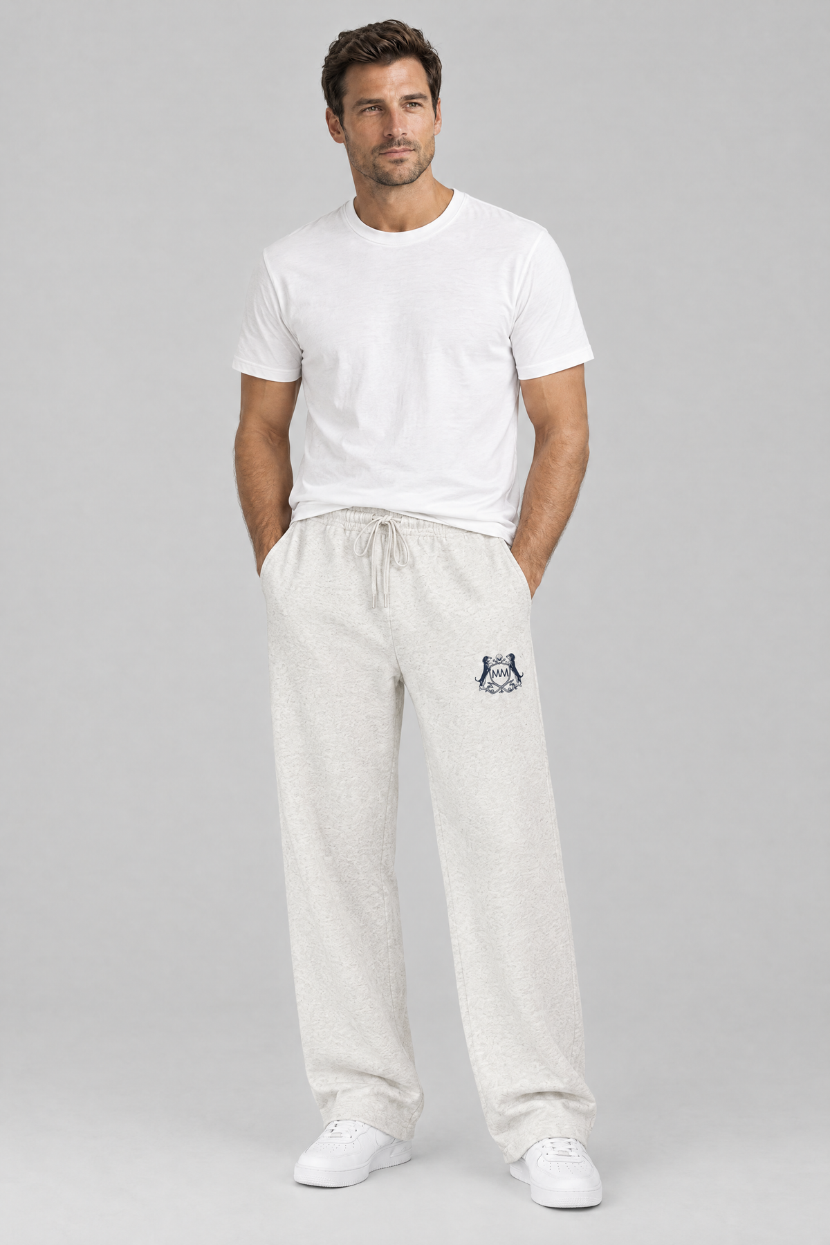 Man standing with his hands in his pockets in oat gray Club Crest sweatpants with the small navy crest and a white tee. Color: Oat Gray