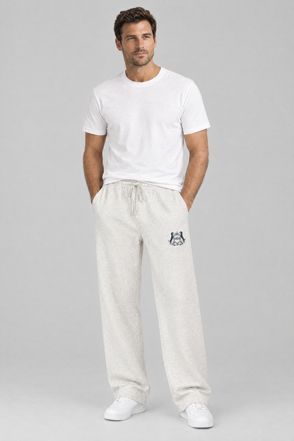 Man standing with his hands in his pockets in oat gray Club Crest sweatpants with the small navy crest and a white tee. Color: Oat Gray