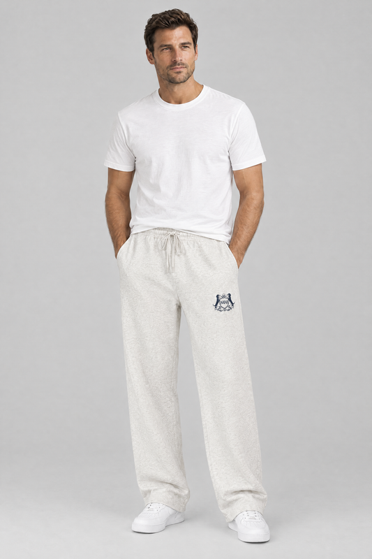 Man standing with his hands in his pockets in oat gray Club Crest sweatpants with the small navy crest and a white tee. Color: Oat Gray