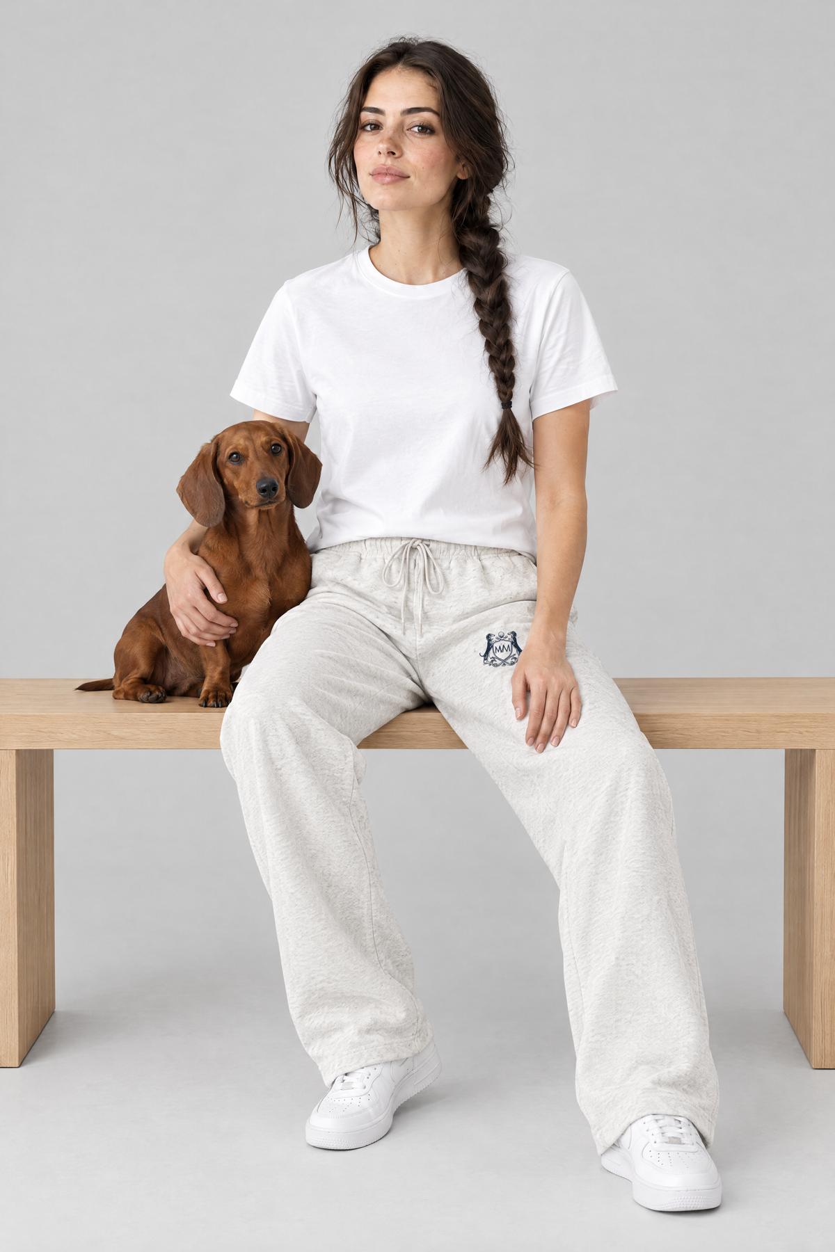 Woman sitting on a wooden bench with a red dachshund, wearing oat gray Club Crest sweatpants and a white tee. Color: Oat Gray
