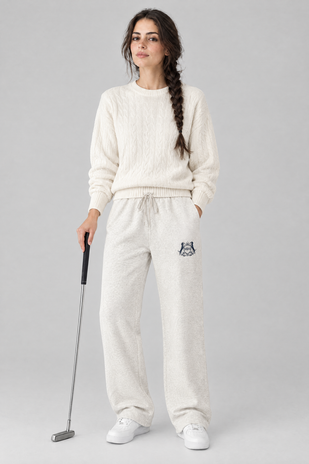 Woman holding a putter in oat gray Club Crest sweatpants with the small navy crest and a cream cable-knit sweater. Color: Oat Gray