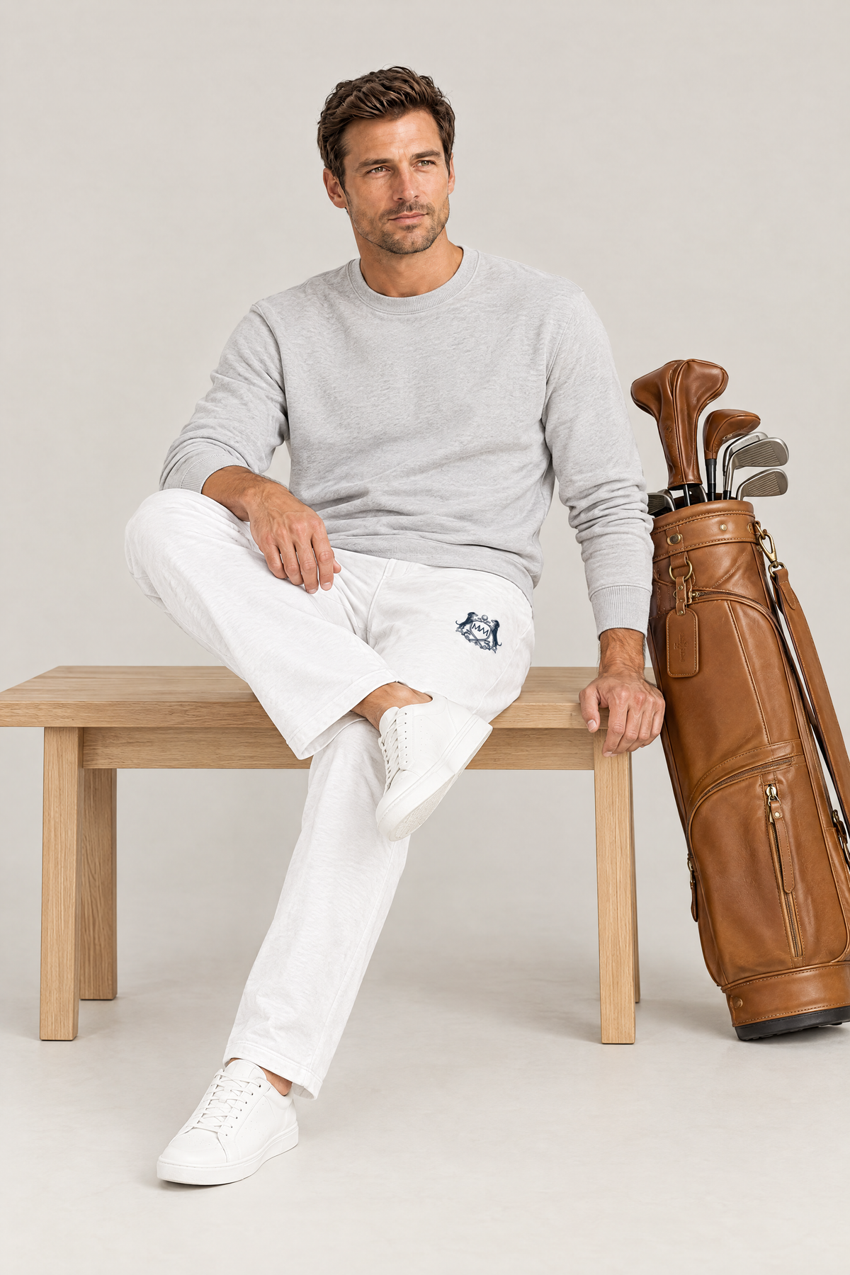 Man sitting on a wooden bench in white Club Crest sweatpants with the small navy crest on the thigh, next to a leather golf bag