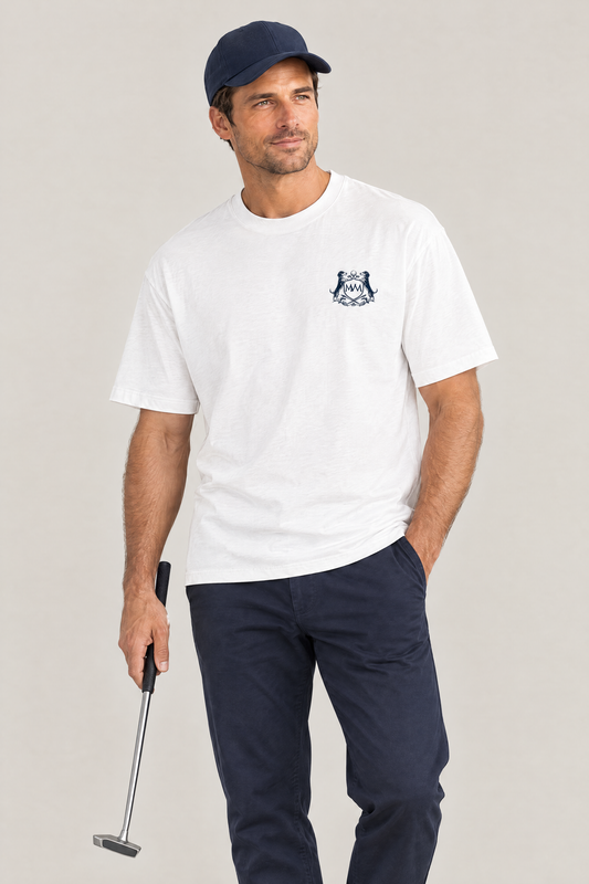 Man holding a putter in the white Club Crest tee with the small navy crest on the chest, navy chinos and a navy cap