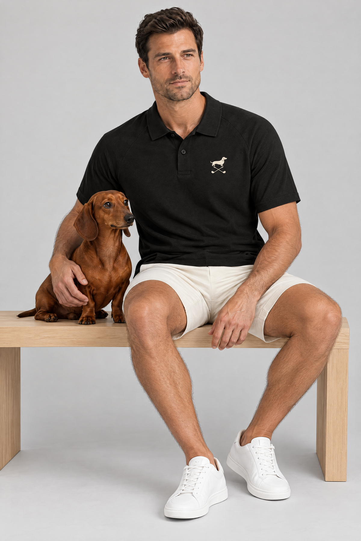 Man sitting on a wooden bench with a red dachshund in the black Club Mark dachshund golf polo and cream golf shorts. Color: Black