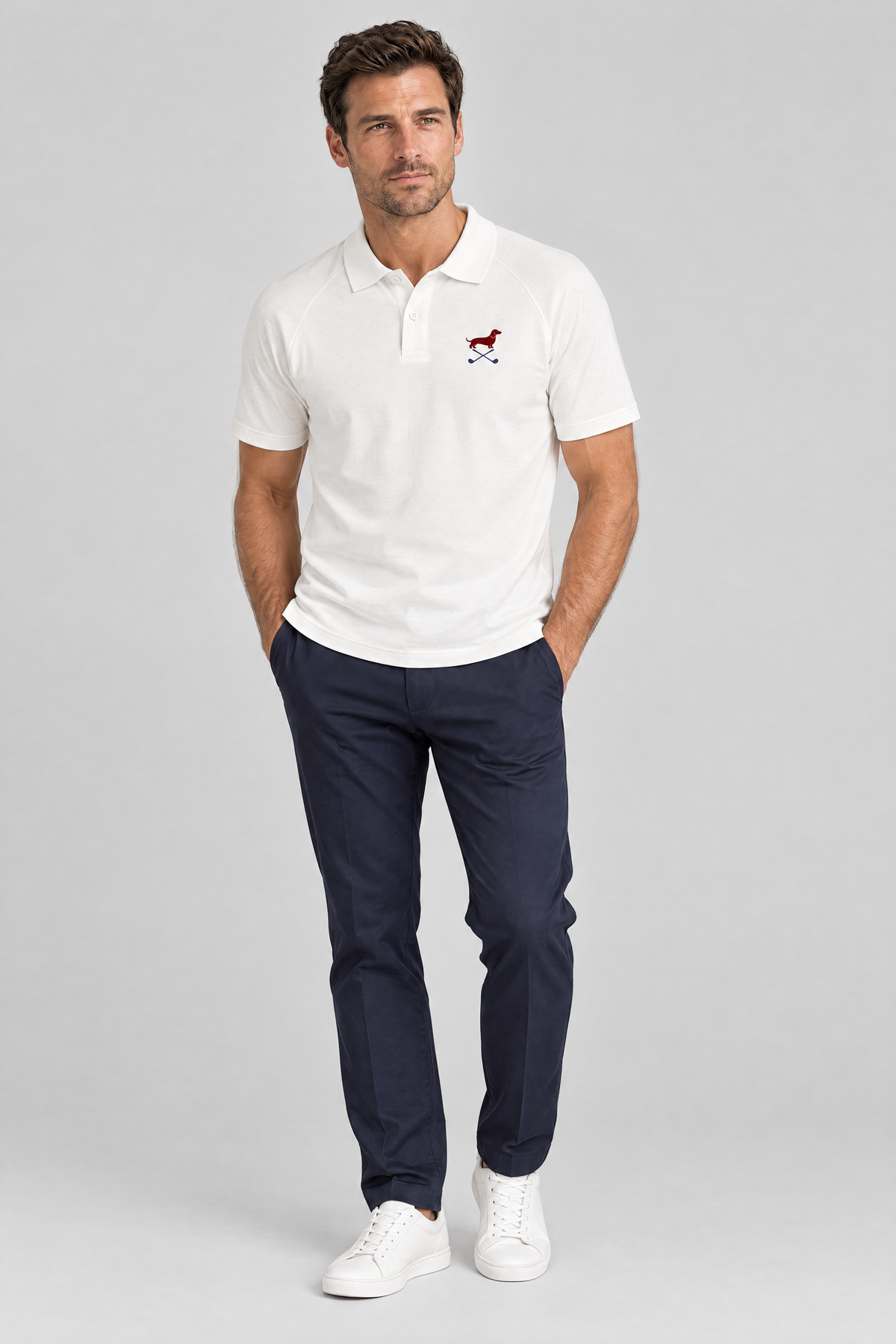 Man standing with his hands in his pockets in the white Club Mark dachshund golf polo and navy trousers. Color: White