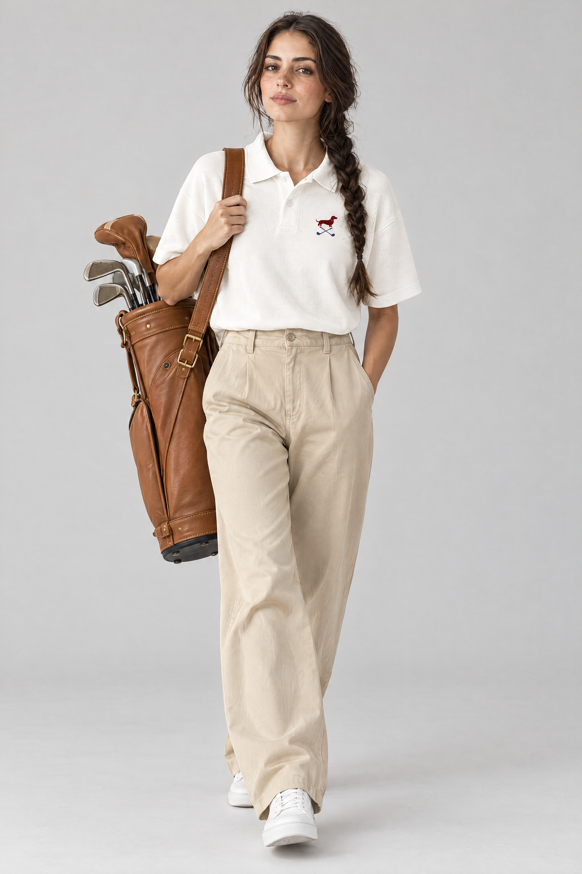 Woman carrying a tan leather golf bag in the white Club Mark dachshund golf polo and beige wide-leg chinos. Color: White
