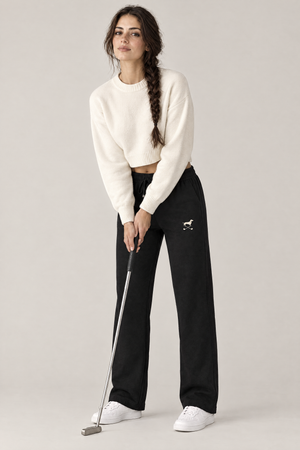 Woman in a relaxed putting stance with a putter in black Club Mark dachshund golf sweatpants with the small cream dachshund and crossed golf clubs logo on the thigh and a cream cropped sweater. Color: Black