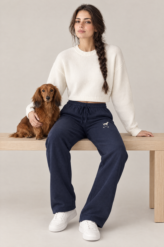 Woman sitting on a wooden bench with a long-haired dachshund in navy blue Club Mark dachshund golf sweatpants with the small cream logo on the thigh and a white cropped sweater. Color: Navy Blue
