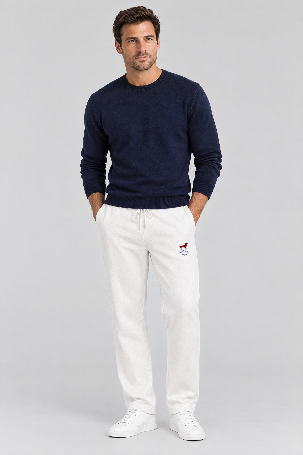 Man standing with his hands in his pockets in white Club Mark dachshund golf sweatpants with the small red dachshund logo and a navy sweater. Color: White