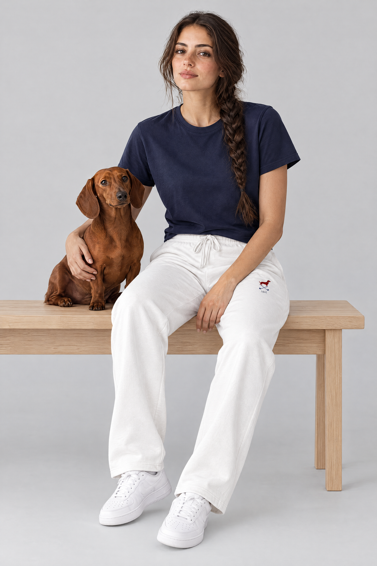 Woman sitting on a wooden bench with a red dachshund, wearing white Club Mark dachshund golf sweatpants and a navy tee. Color: White