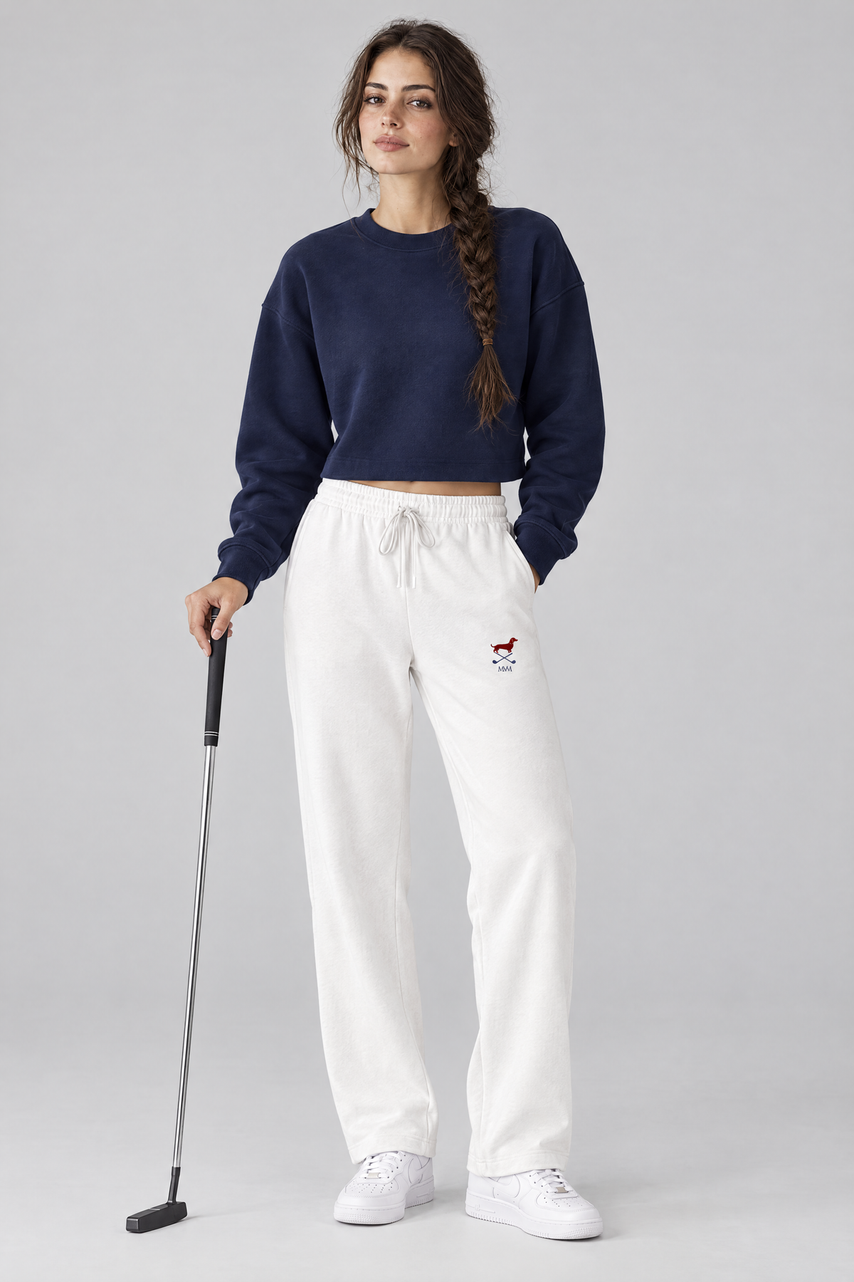 Woman holding a putter in white Club Mark dachshund golf sweatpants with the small red dachshund and crossed golf clubs logo on the thigh and a navy cropped sweater. Color: White