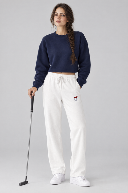 Woman holding a putter in white Club Mark dachshund golf sweatpants with the small red dachshund and crossed golf clubs logo on the thigh and a navy cropped sweater. Color: White