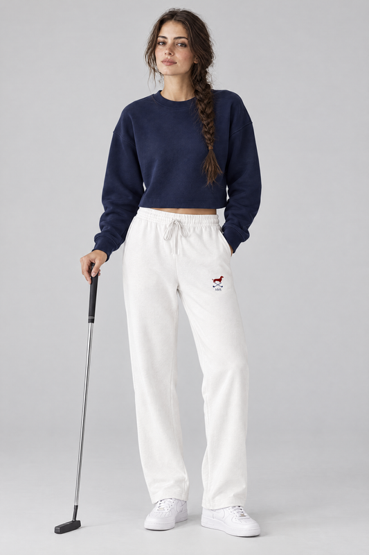 Woman holding a putter in white Club Mark dachshund golf sweatpants with the small red dachshund and crossed golf clubs logo on the thigh and a navy cropped sweater. Color: White