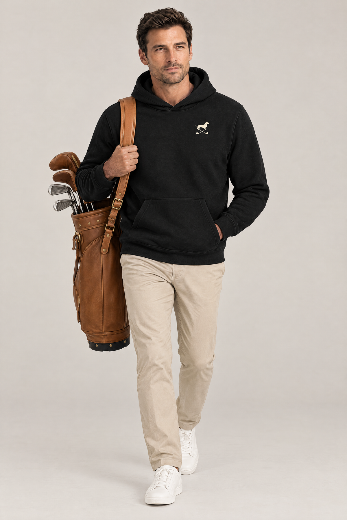 Man carrying a tan leather golf bag in the black Club & Swing dachshund golf hoodie and stone chinos. Color: Black