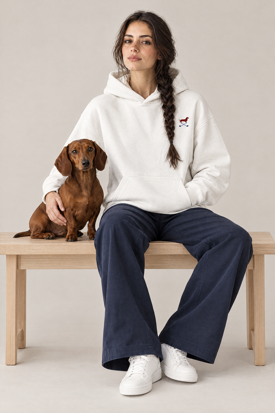 Woman sitting on a wooden bench with a red dachshund in the white Club & Swing dachshund golf hoodie and navy wide-leg trousers. Color: White