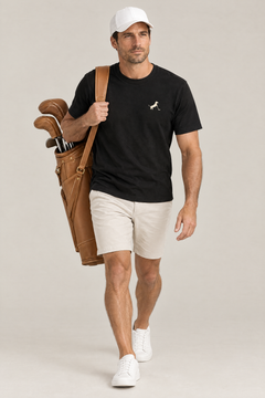 Man walking with a tan leather golf bag over his shoulder in the black Putting dachshund golf t-shirt, stone chino shorts and a white cap. Color: Black