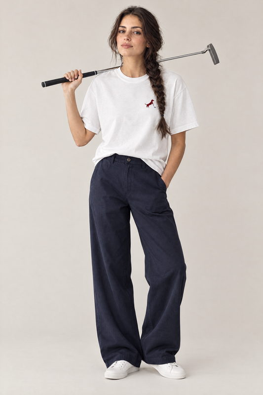 Woman holding a putter over her shoulder in the white Putting dachshund golf t-shirt and navy wide-leg trousers. Color: White
