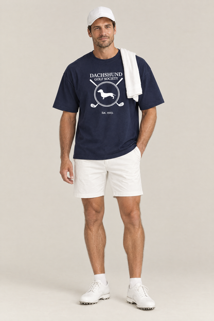 Man wearing the Dachshund Golf Society Tee in navy with white golf shorts and a towel over his shoulder. Color: Navy