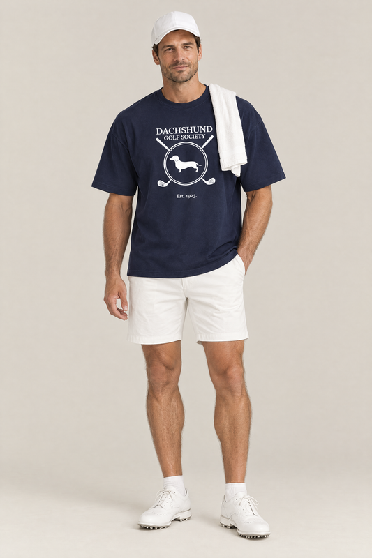 Man wearing the Dachshund Golf Society Tee in navy with white golf shorts and a towel over his shoulder. Color: Navy